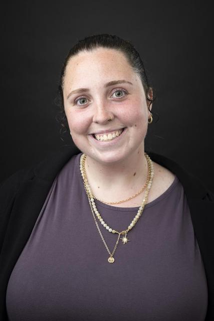 Professional headshot of person in purple blouse with black sweater against a black background.